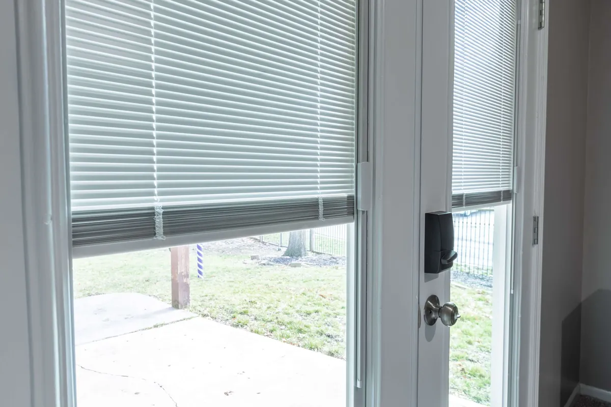 This interior image features a close-up of blinds, a door handle, and an outdoor view through the glass. The scene is bright, airy, and calm, suggesting the transition from outside to inside while offering a glimpse of the serene neighborhood beyond.