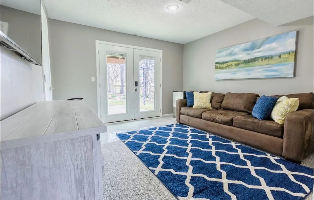 A bright, welcoming living room featuring a cozy sofa, a textured rug, stylish wall art, and sunlit double doors that open to the outside. The space feels airy and inviting, perfect for both relaxing and socializing.