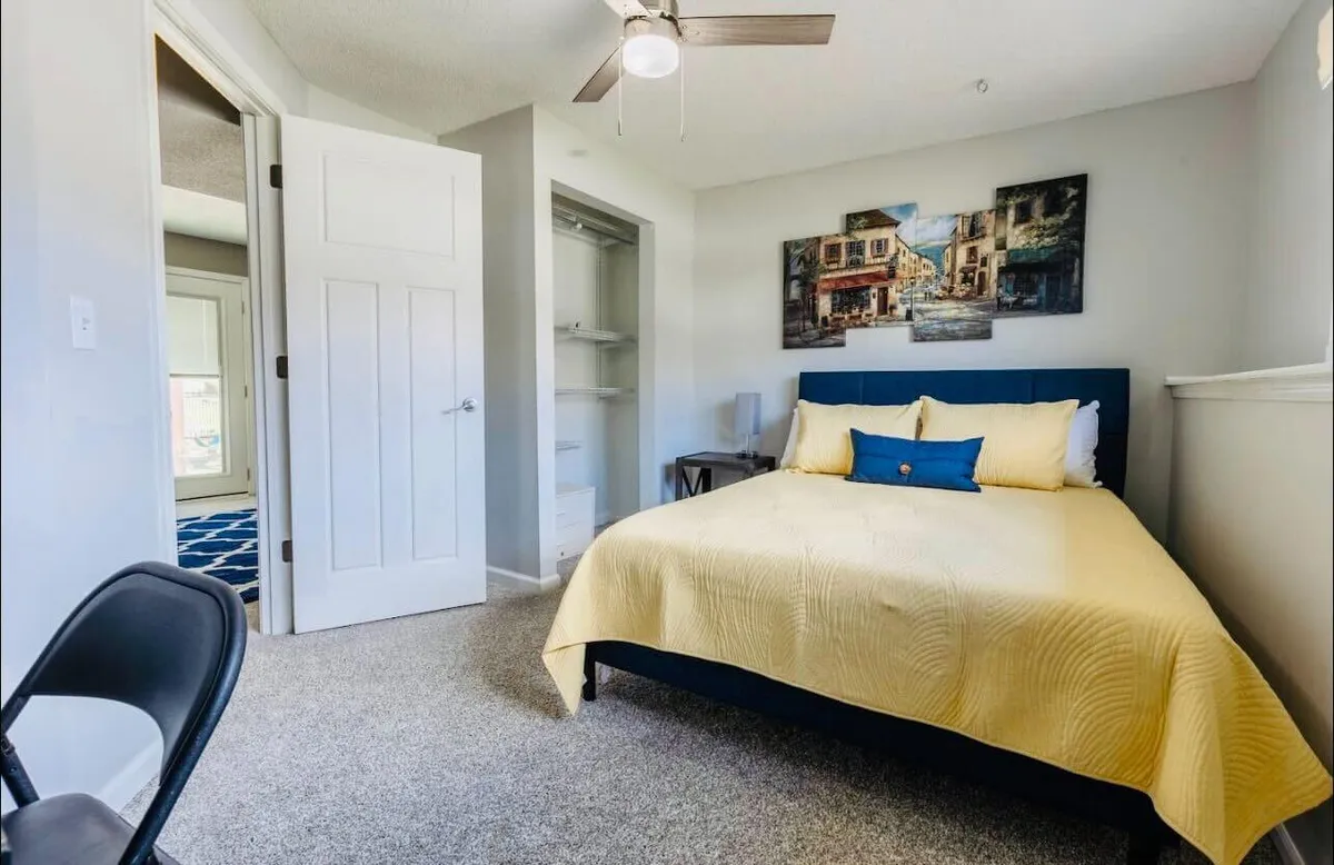 An inviting bedroom scene with a bed draped in a yellow blanket, framed artwork on the wall, and a chair next to the bed. An open door leads to another room, suggesting an easy flow between spaces and the potential proximity to the kitchen area. The mood is cozy, ideal for unwinding after a long day or enjoying quiet moments.